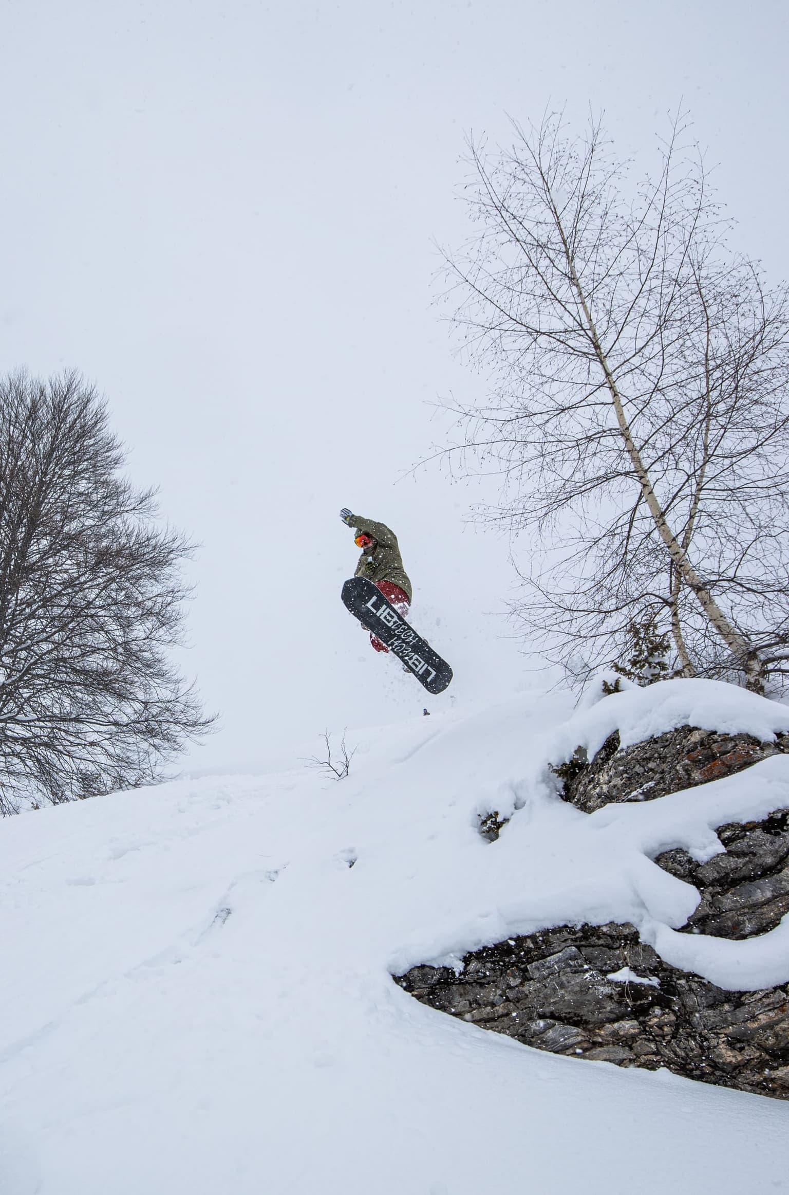 Snowboarder launching off a snowy feature beside a rocky drop.